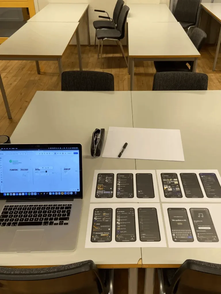 A computer on a table and paper prototypes of a Spotify playlist to start the research